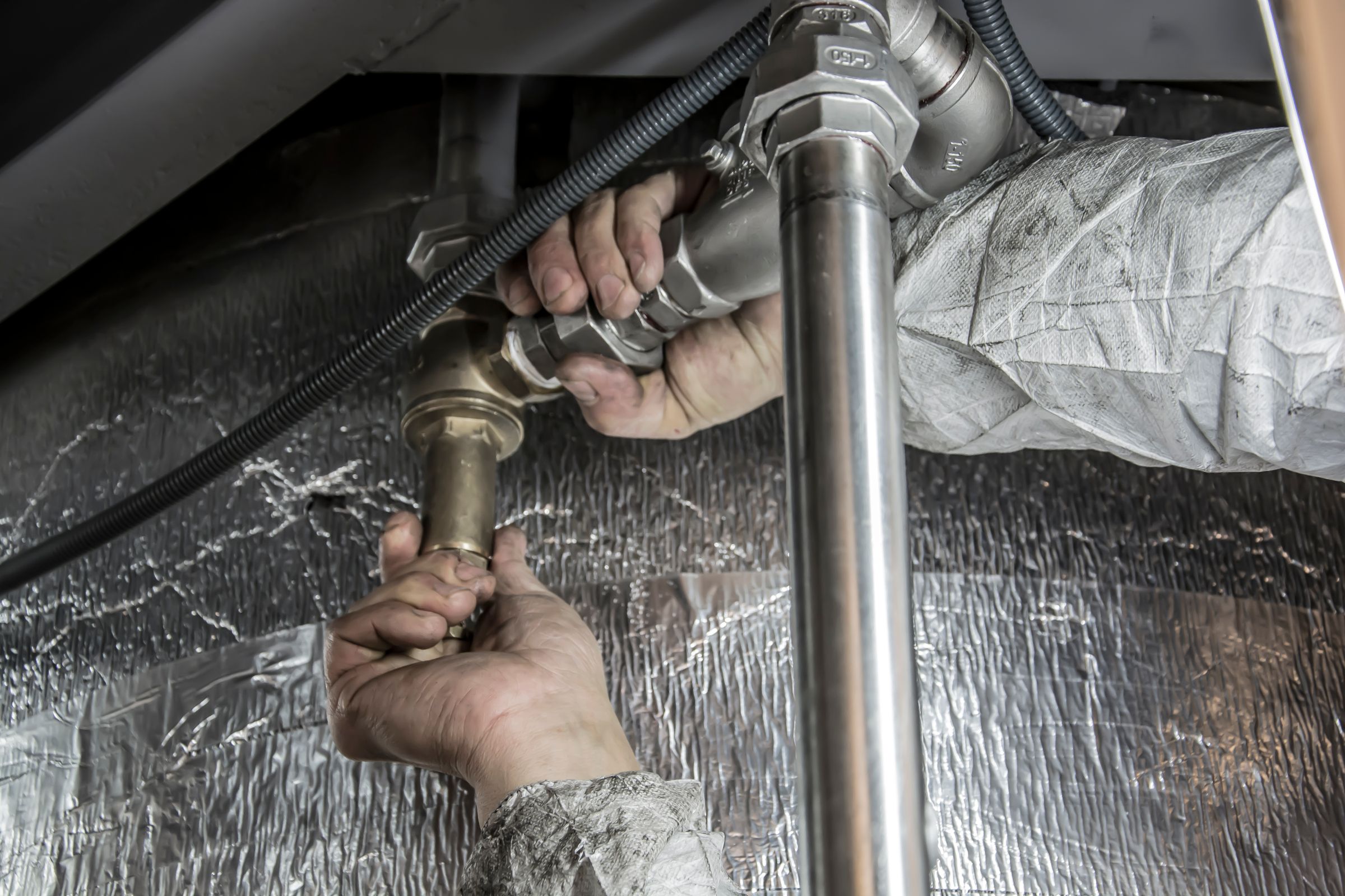 Plumber's hands tightening pipe fittings in a cramped utility space with foil insulation behind the work. Photo by Anıl Karakaya on Pexels.