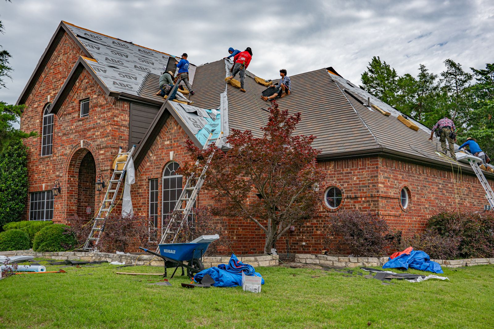 Roofing crew replacing shingles on a brick house, with ladders, underlayment, and a wheelbarrow on the lawn. Photo by Ryan Stephens on Pexels.