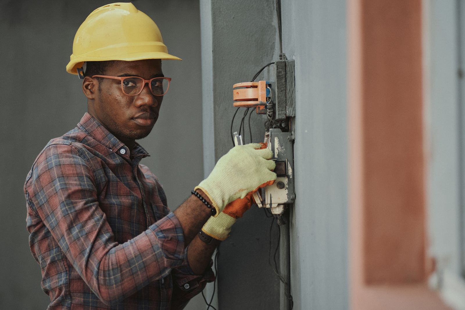 Electrician in a yellow hard hat and gloves working on a wall-mounted fuse box. Photo by Emmanuel Ikwuegbu on Pexels.